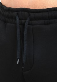 Detailní záběr na černý elastický pás se šňůrkami na černých teplácích, které jsou proti viditelné kůži.