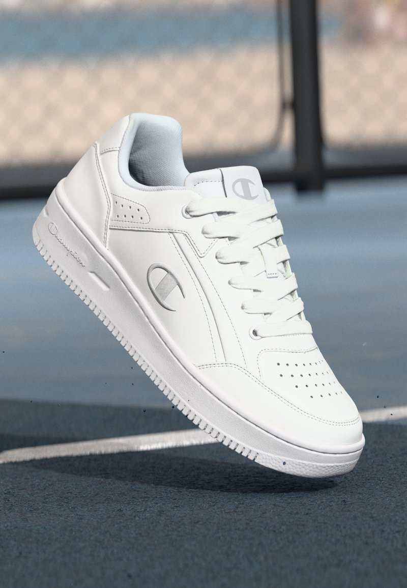 White leather sneaker with flat laces, perforated toe, and chunky sole. Features a subtle logo and smooth texture. Suitable for casual wear.