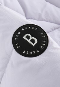 Fehér steppelt anyag, amelyen egy kör alakú fekete jelvény található "TED BAKER" felirattal és egy kiemelkedő ezüst "B" logóval. Simák és könnyű tapintású.