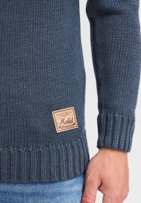 Marineblauer Strickpullover mit gerippten Bündchen und Saum. Mit einem hellbraunen Lederpatch mit geprägt Text "Solid" und Markendetails auf der linken Seite.