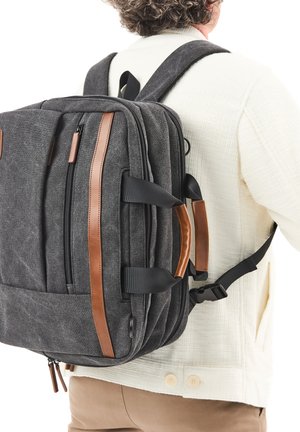 Person, der einen dunkelgrauen Rucksack mit braunen Akzenten und mehreren Reißverschlussfächern über einer weißen Jacke und beigen Hosen trägt.