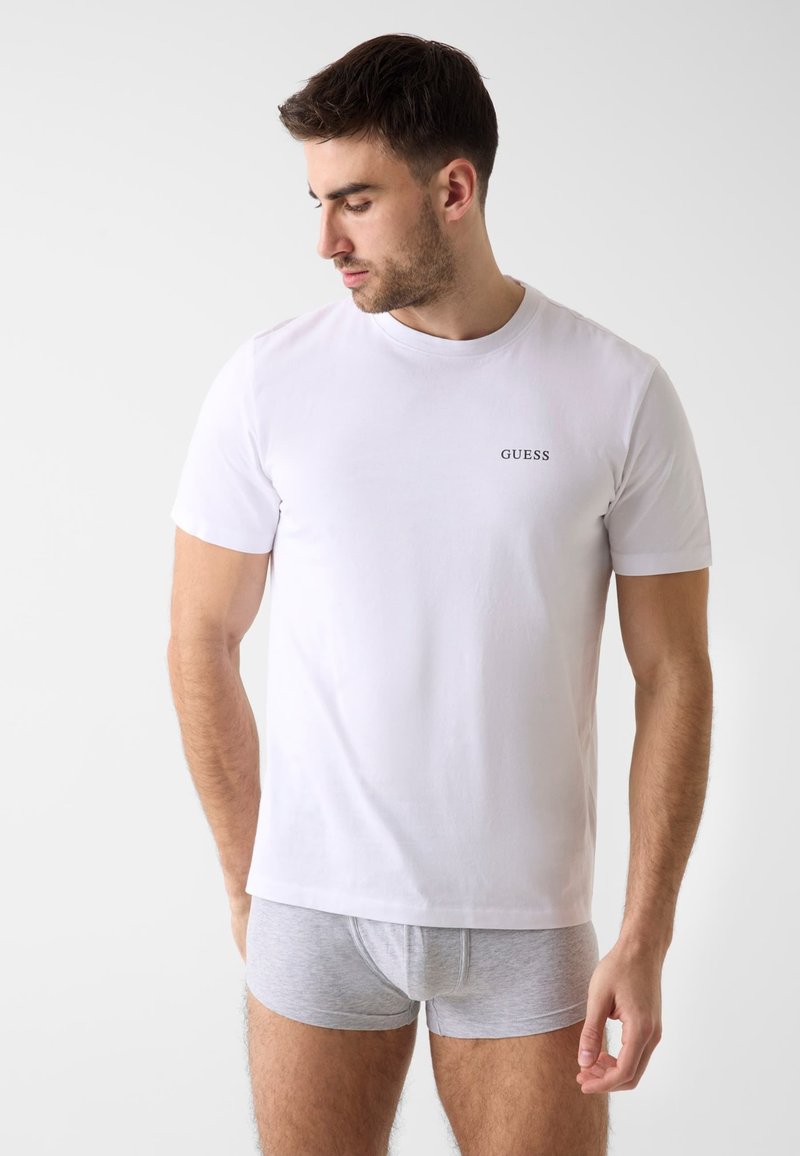 Homme portant un t-shirt blanc Guess et un boxer gris clair, debout devant un fond blanc uni, regardant vers le bas à sa gauche.