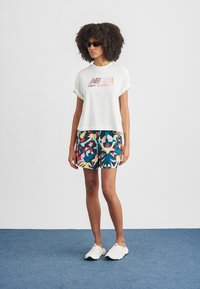 T-shirt branca curta com logótipo "New Balance", combinada com calções coloridos estampados; modelo a usar óculos de sol pretos e calçado branco.
