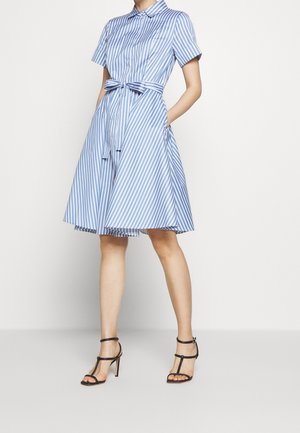 Vrouw die een blauw-wit gestreepte jurk met korte mouwen en een taillekoord draagt, staand met handen in de zakken en zwarte sandaalhakken.