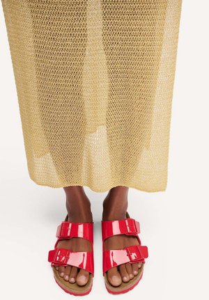 Voeten met glanzende rode sandalen met dubbele band onder een beige gebreide rok, op een lichte achtergrond.