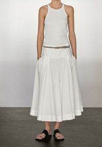 Jupe plissée blanche avec un ourlet large, associée à un débardeur blanc ajusté et une ceinture beige ornée de clous en métal, portée avec des sandales noires.