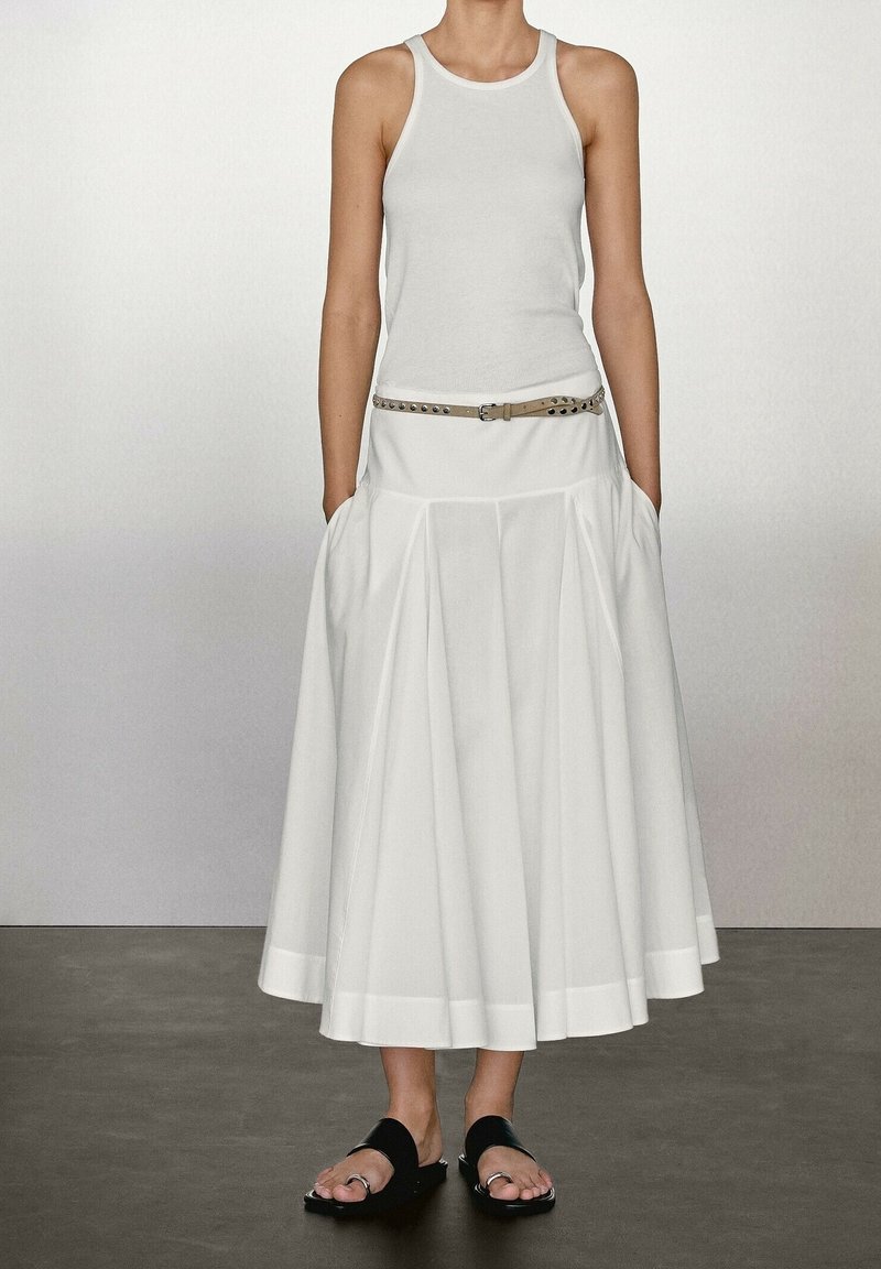 Jupe plissée blanche avec un ourlet large, associée à un débardeur blanc ajusté et une ceinture beige ornée de clous en métal, portée avec des sandales noires.