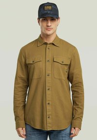 Camicia verde oliva con bottoni, dotata di due tasche sul petto, maniche lunghe e orlo dritto. Indossata con un cappellino nero mimetico con una patch del logo.