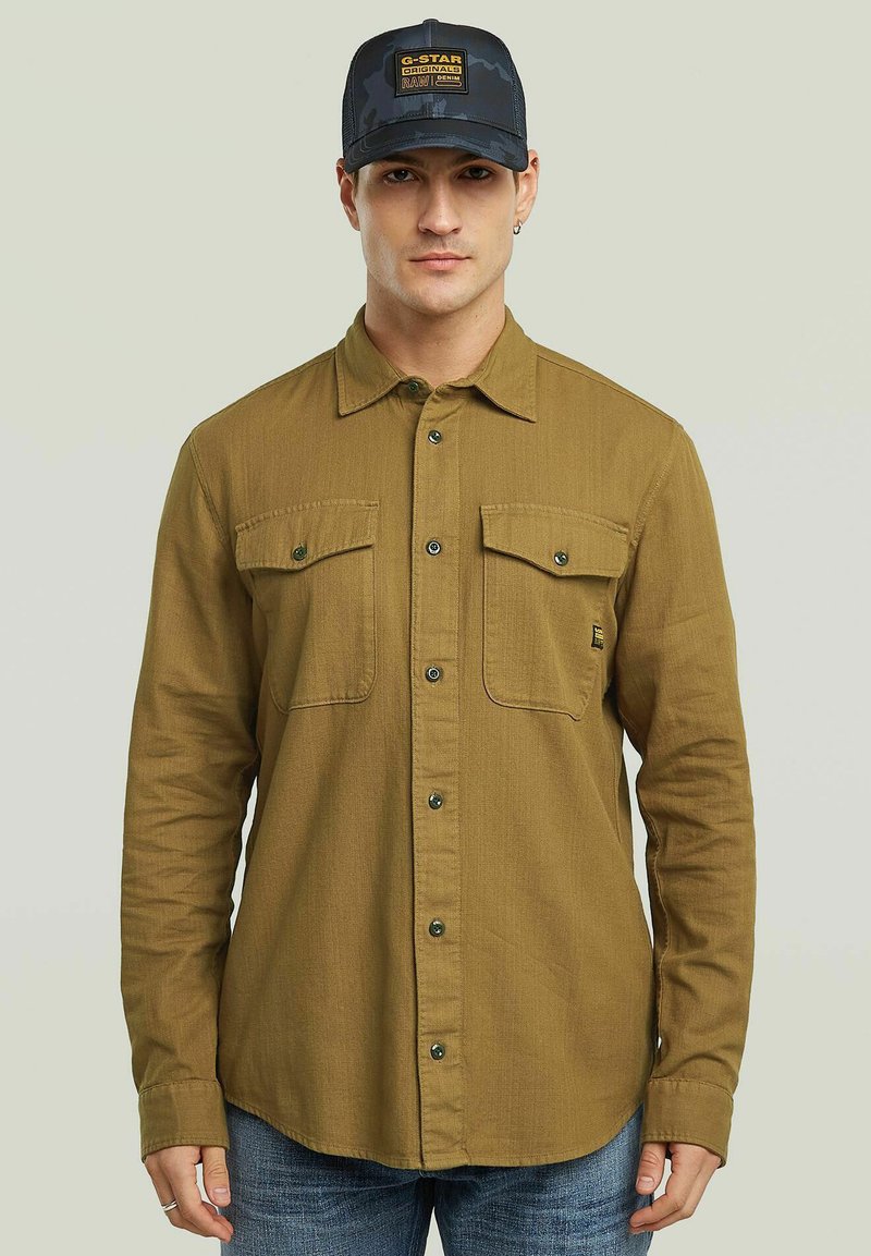 Camicia verde oliva con bottoni, dotata di due tasche sul petto, maniche lunghe e orlo dritto. Indossata con un cappellino nero mimetico con una patch del logo.