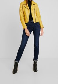 Giubbotto biker in pelle gialla con zip asimmetrica, abbinato a jeans skinny blu scuro e stivali neri alla caviglia. Texture liscia e design aderente.