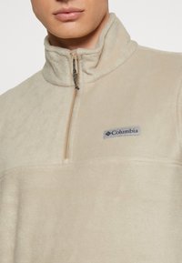 Beżowy polarowy sweter z wysokim kołnierzem, pół-zamkiem z przodu i naszywką z logo Columbia. Miękka faktura i subtelny połysk.