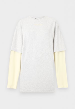 Vrouw met een beige hoed met zwart ontwerp, gelaagde oversized witte en lichtgele tops, ruime blauwe spijkerbroek en zwarte schoenen, staand met gekruiste armen.