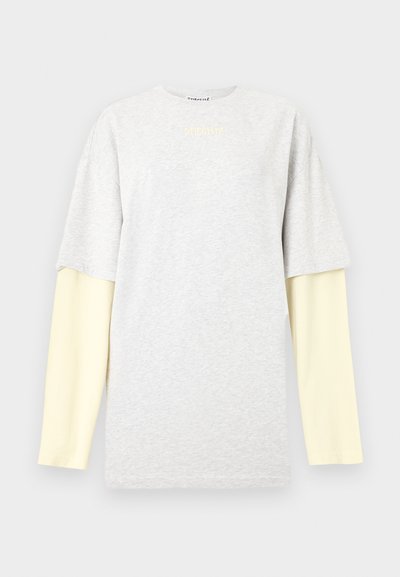 Ανοιχτό γκρι κοντομάνικο πουκάμισο φορεμένο πάνω από μακριά, αχνά κίτρινα μανίκια, με μικρό λογότυπο κειμένου στο κέντρο κοντά στο λαιμό.