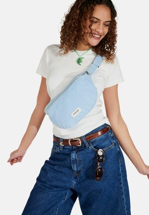 Femme souriante portant un t-shirt blanc, un jean bleu, une ceinture marron, un collier poisson vert, un sac bandoulière bleu clair et des lunettes de soleil accrochées au jean.