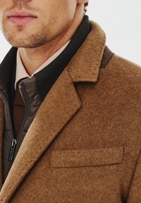 Brązowy płaszcz z mieszanki wełny z wyciętym kołnierzem, nałożony na ciemną kurtkę puchową. Posiada kieszeń na piersi oraz teksturowany materiał.