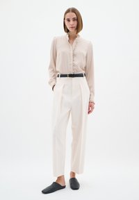 Blusa crema con taschino arricciato, abbinata a pantaloni a pieghe bianchi a vita alta e scarpe nere a scivolo. Design minimalista e vestibilità sartoriale.