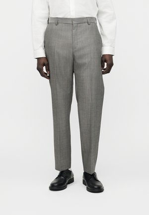 Férfi szürke szabott nadrágban, fehér ingben és fekete elegáns cipőben, egyszerű fehér háttér előtt áll.