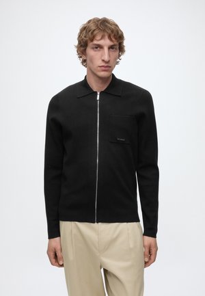 Young man with wavy blond hair wears a black zip-up sweater with a chest pocket and beige pleated trousers against a plain background.