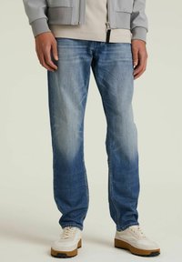 Vaqueros de mezclilla azul claro con un acabado desgastado, corte recto, diseño de cinco bolsillos y costuras visibles, combinados con zapatillas de color claro.