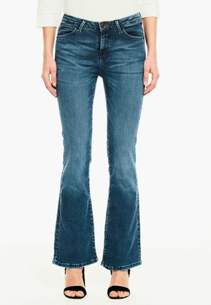 Jean flare en denim délavé foncé avec une taille ajustée, ourlet effiloché et coutures contrastantes. Associé avec des talons ouverts noirs.