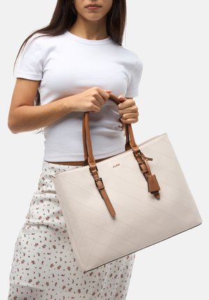 Beige geborduurde draagtas metbruine leren handvaten en details, met een gestructureerde vorm en minimale hardware-accenten.