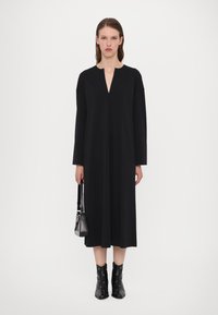 Black long-sleeve dress with a V-neckline; smooth fabric, loose fit, and knee-length design. Model holds a small black handbag.