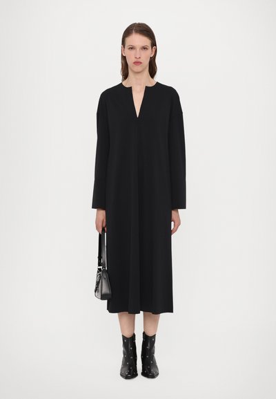 Crna haljina dugih rukava s V-izrezom; glatka tkanina, slobodan kroj i dizajn do koljena. Manekenka drži malu crnu torbicu.