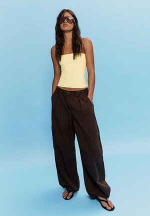 Młoda kobieta o długich ciemnych włosach, nosząca duże okulary przeciwsłoneczne, blady żółty top bez ramiączek, luźne ciemnobrązowe spodnie i czarne klapki, stojąca.