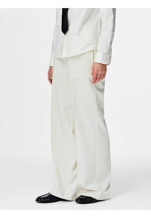 Person, die weiße, breitbeinige Hose mit Nadelstreifen, weißes Hemd, schwarze Krawatte und schwarze flache Schuhe trägt, steht vor einem schlichten Hintergrund.