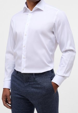 Homme portant une chemise blanche ajustée à manches longues rentrée dans un pantalon texturé gris foncé, avec une main dans la poche.