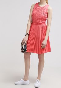 Plisowana sukienka letnia w kolorze koralowym, z teksturowanym górnym częścią, bez rękawów oraz rozkloszowaną spódnicą. Stylizowana z białymi trampkami i kwiecistą kopertówką.