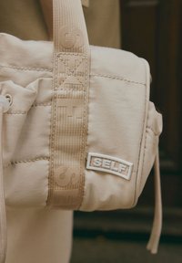 Beige olka-käerotasku, jossa on pehmustettu pinta. Tunnusomaista on leveä olkahihna, jossa on kohokuvioitu "SELF" -teksti ja suorakulmainen logopatch.
