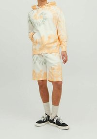 Hoodie w stylu tie-dye w białych, pomarańczowych i zielonych odcieniach, z kieszenią z przodu; dopasowane szorty w stylu tie-dye; połączone z czarnymi trampkami i białymi skarpetkami.