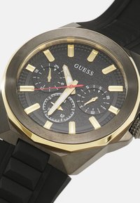 Montre Guess pour homme avec un cadran noir texturé, index en or, aiguille des secondes rouge et bracelet en caoutchouc. Dispose de sous-cadrans pour la date et l'heure.