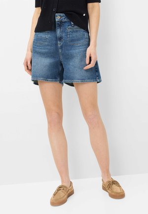 Frau trägt blaue Jeansshorts, schwarzen strukturierten Top und hellbraune Schnürschuhe, steht vor weißem Hintergrund.