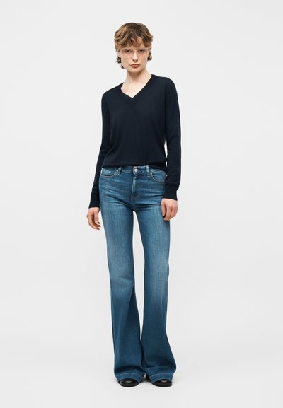 Jeune personne portant des lunettes transparentes, un haut noir à manches longues avec un col en V, et un jean bleu évasé taille haute, debout devant un fond uni.