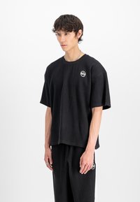 Ung mann med kort, kr�øllete hår iført en svart, teksturert oversized t-skjorte og matchende bukser med hvite logopatcher, står mot en hvit bakgrunn.