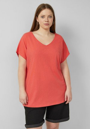 Młoda kobieta stoi, ubrana w luźny koralowy T-shirt z dekoltem w serek i czarne podwinięte szorty na jednolitym szarym tle.