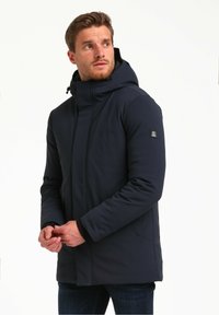 Chaqueta impermeable de color navy con capucha, cremallera frontal y dos bolsillos laterales. Presenta un acabado suave y un parche con el logotipo en la manga izquierda.