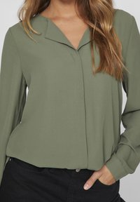Blusa verde oliva con scollatura a V e maniche lunghe, caratterizzata da una texture morbida e cuciture sottili, abbinata a pantaloni neri.