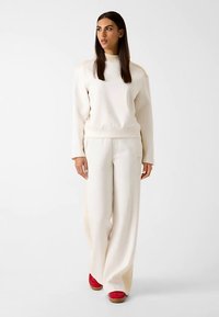 Cream sweatshirt with ribbed collar and long sleeves paired with wide-leg cream pants featuring textured side details. Red shoes complete the outfit.