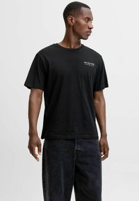 Hombre vestido con una camiseta negra de manga corta con texto blanco en el lado izquierdo del pecho y vaqueros oscuros holgados, de pie contra un fondo claro y liso.