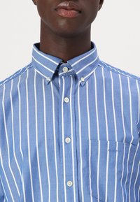 Blauwe shirt met witte verticale strepen, button-down kraag, een voorzak en twee knopen bij de kraag. Katoenen materiaal, gladde textuur.