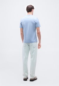 Νεαρός άνδρας στέκεται με την πλάτη προς το μέρος μας, φορώντας ανοιχτό μπλε κοντομάνικο πουκάμισο, ανοιχτό μπλε τζιν και μπεζ παπούτσια σε απλό λευκό φόντο.