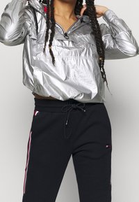 Silverfärgad, metallisk anorak med dragkedja fram, elastiska ärmmuddar och ficka. Svarta sweatpants med sidostripes i rött, blått och vitt.