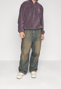 Fialový fleecový pulover s čtvrtkovým zipem a vyšitým logem, spárovaný se světle modrými džínovými cargo kalhotami s bočními kapsami a vybledlým vzhledem.