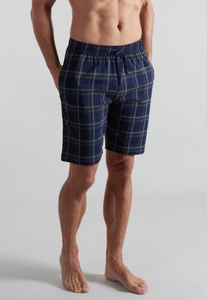 Man wearing navy blue plaid knee-length shorts with elastic waistband and drawstring, standing barefoot against plain background.