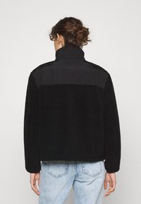 Czarna kurtka z futra syntetycznego z wysokim kołnierzem oraz gładkimi akcentami na rękawach i ramionach, o luźnym kroju i prostym wzornictwie.