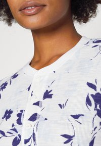 Un pull blanc à boutons présentant un tissu texturé, des motifs de fleurs violettes et un col rond avec quatre boutons blancs.