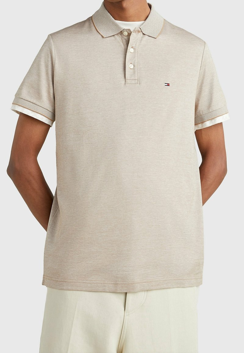 Polo beige à manches courtes avec trois boutons et petit logo sur la poitrine, porté par-dessus un sous-vêtement blanc avec poignets visibles.
